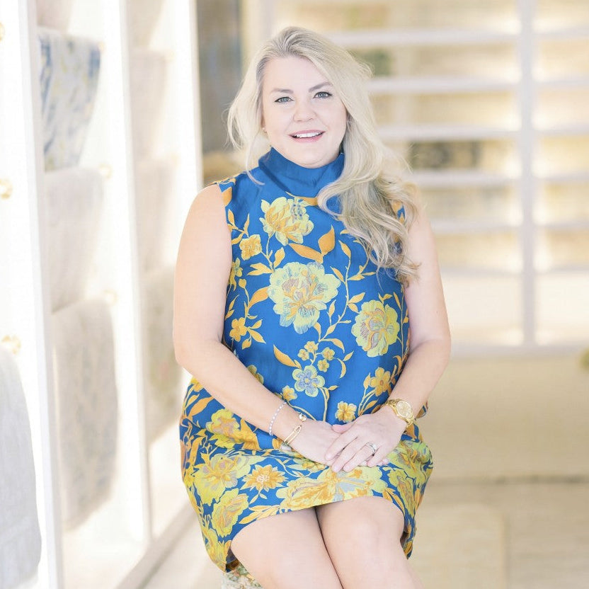 Woman in a blue and yellow floral dress sitting in a bright room with white walls and furniture.